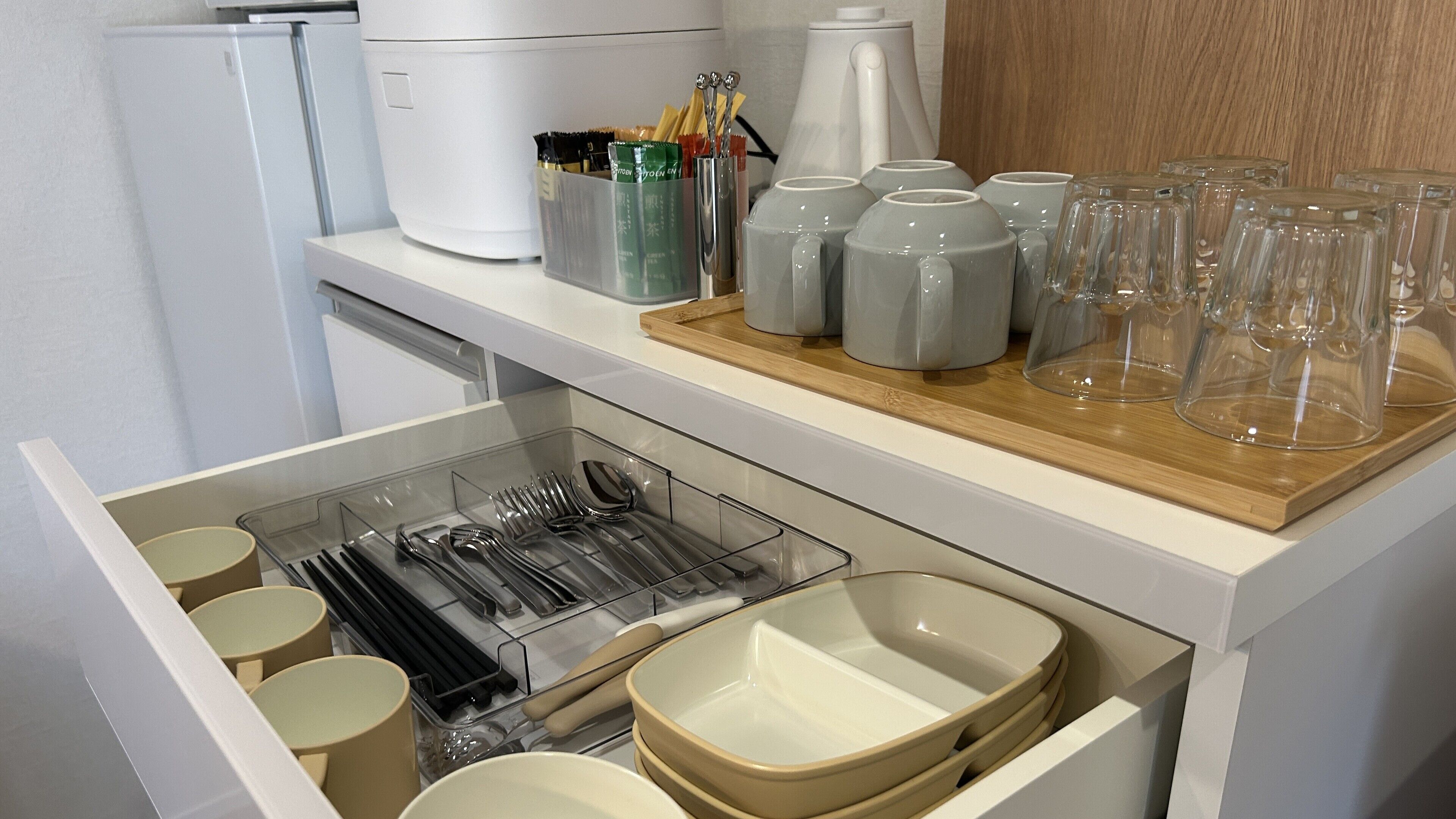 A kitchen with a drawer containing utensils and bowls, a coffee maker, and a tray with glasses and cups.