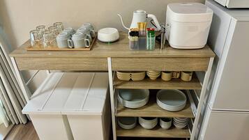 A kitchen area with a wooden countertop, a coffee maker, a tea kettle, and a tray of glasses. Below, there are shelves with plates and bowls.