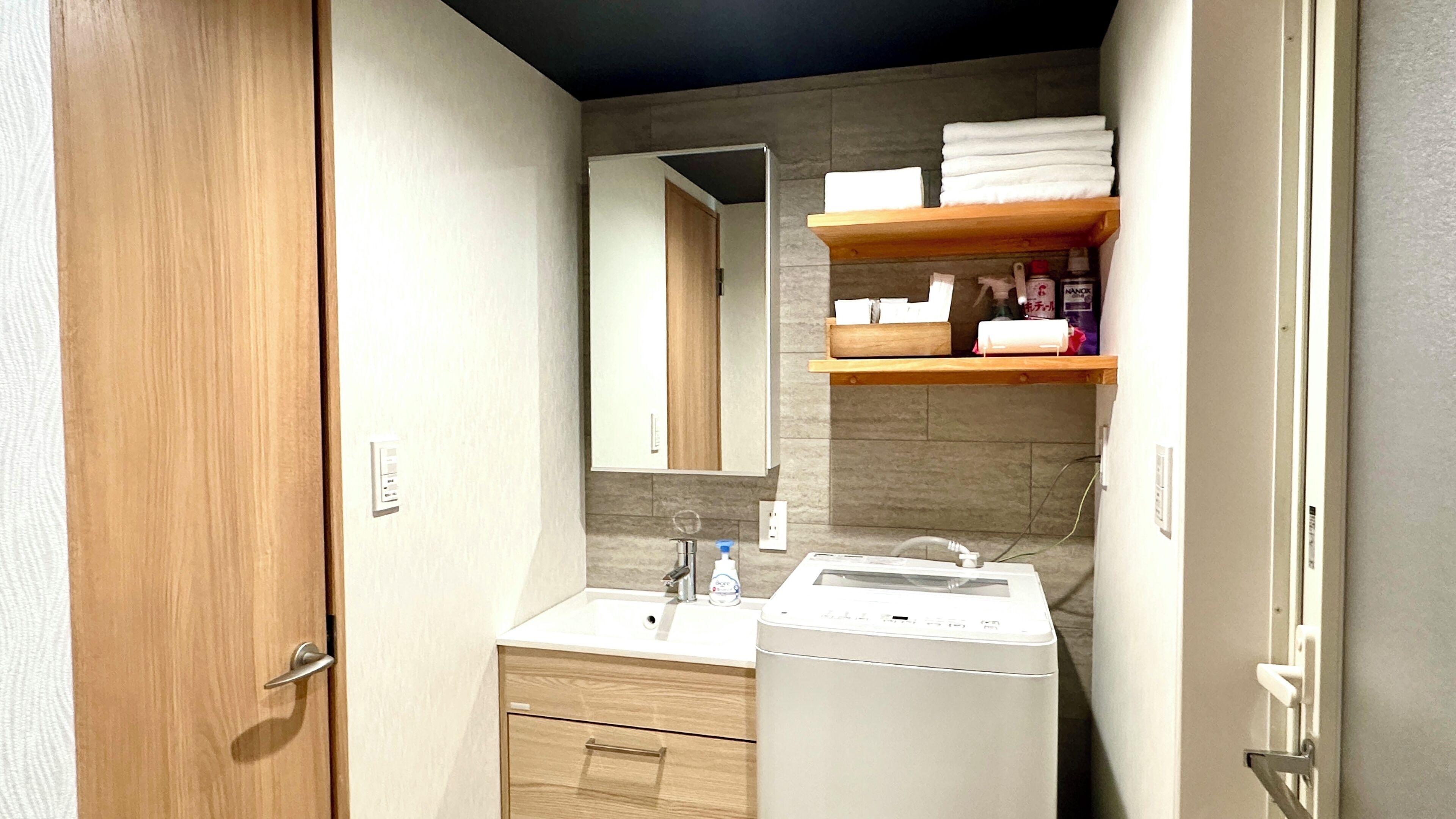 A compact bathroom with a wooden door, a white sink, a mirror, a towel rack, and a washing machine.