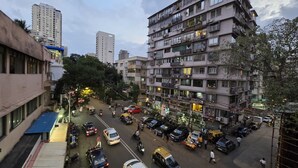 Un immeuble à plusieurs étages, doté de balcons et de climatiseurs, entouré d’autres bâtiments et bordé par une rue animée où circulent des véhicules et des piétons.