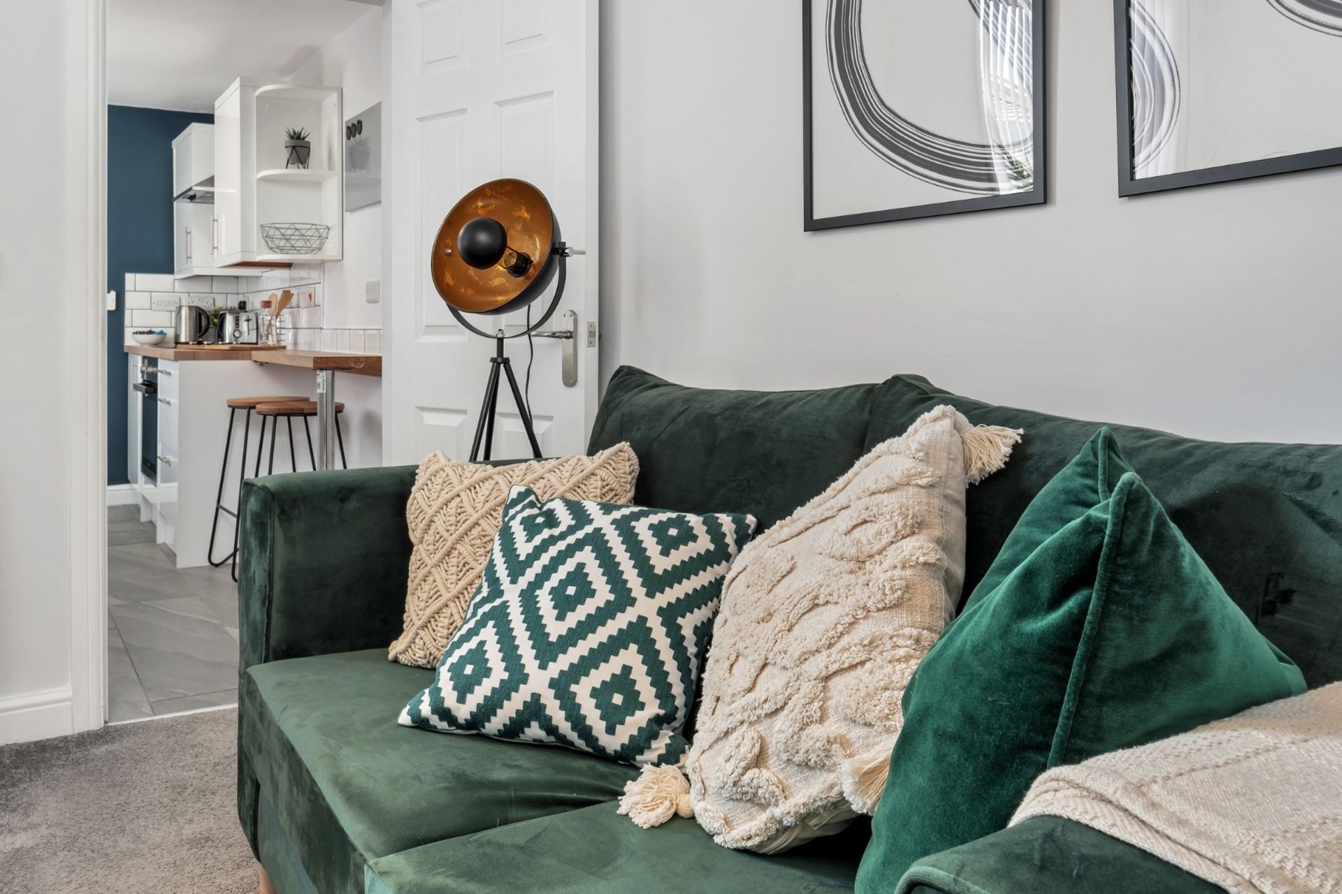 Ein grünes Sofa mit gemusterten Kissen, eine Stehlampe und im Hintergrund ein Küchenbereich.