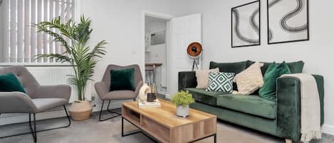 A modern living room with a grey sofa, a wooden coffee table, and a green armchair. There are two abstract paintings on the wall and a potted plant.