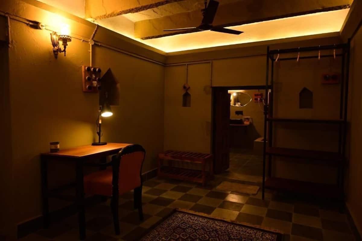 Een kamer met weinig licht, met een houten tafel en stoel, een plafondventilator en een geruit vloertje.