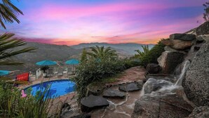 Zona de piscina con cascada, rodeada de rocas y vegetación, y vistas a montañas distantes bajo un cielo colorido.