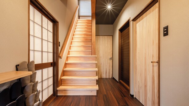 Una escalera de madera en un pasillo con una lámpara en el techo, puertas de madera y una zona de estar con sillas.