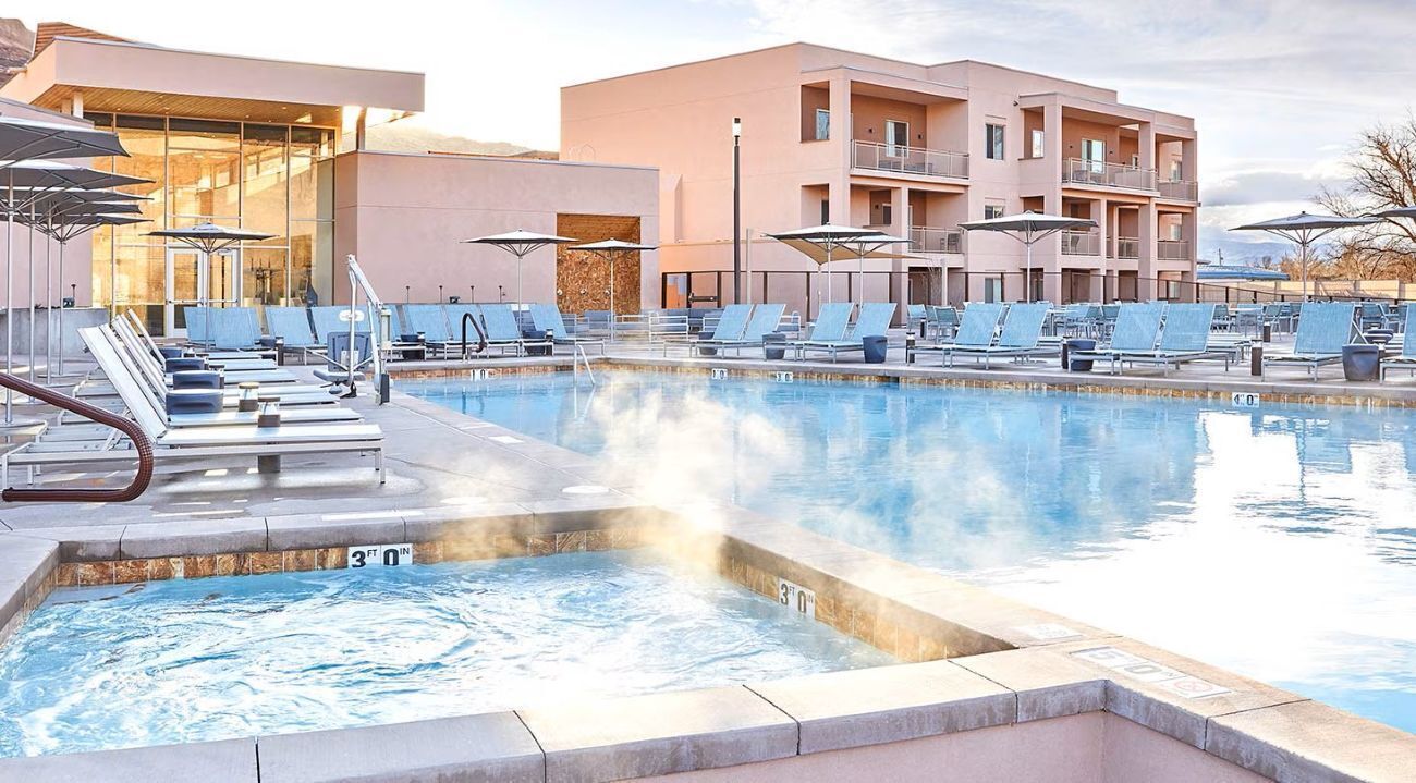 A swimming pool with lounge chairs and umbrellas, a hot tub, and a modern building with balconies.