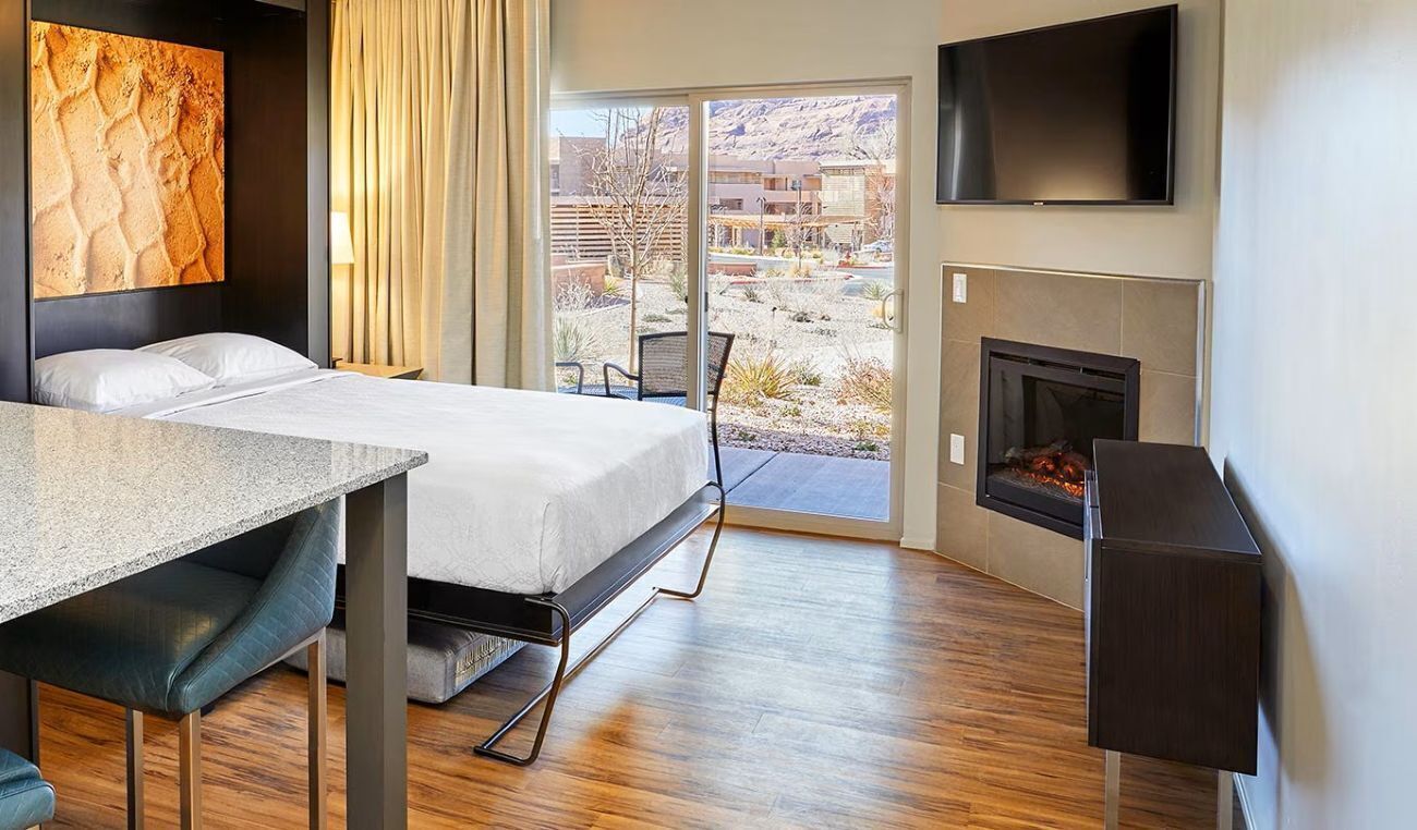 Una habitación de hotel con cama, una mesa de comedor, televisión, chimenea y vistas al paisaje desértico.