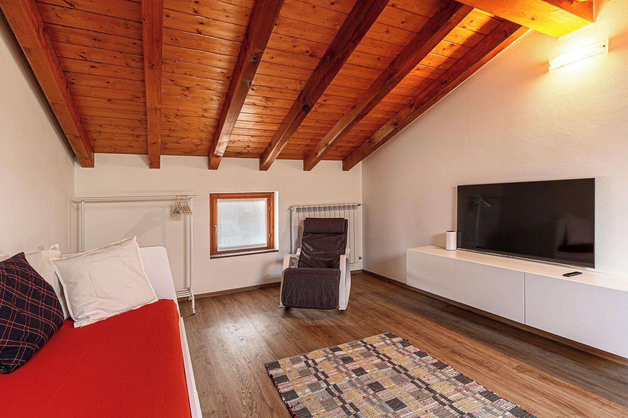 A cozy room with a wooden ceiling, a bed with red bedding, a TV on a white cabinet, and a window with blinds.