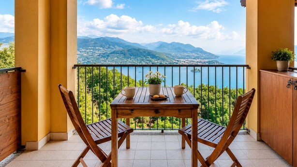 Ein Balkon mit einem Holztisch, zwei Stühlen, einem Topfpflanzen und Blick auf eine gebirgige Landschaft.