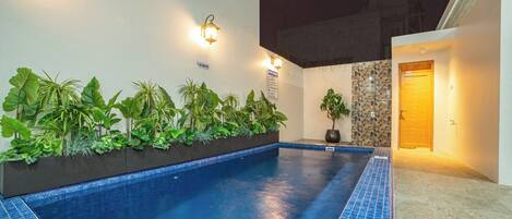 A swimming pool with a tiled edge, surrounded by a planter with tropical plants, a 'No Smoking' sign, and a door leading to a building.