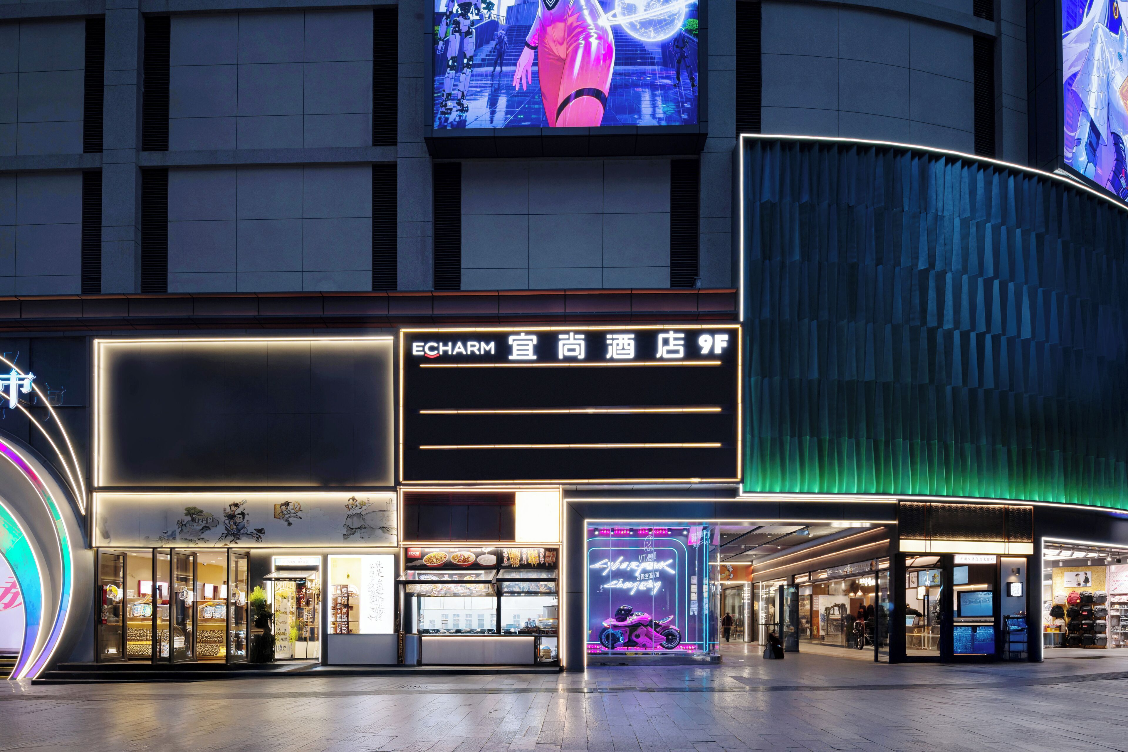 A modern hotel facade with illuminated signage, a digital display, and a storefront with visible food items.