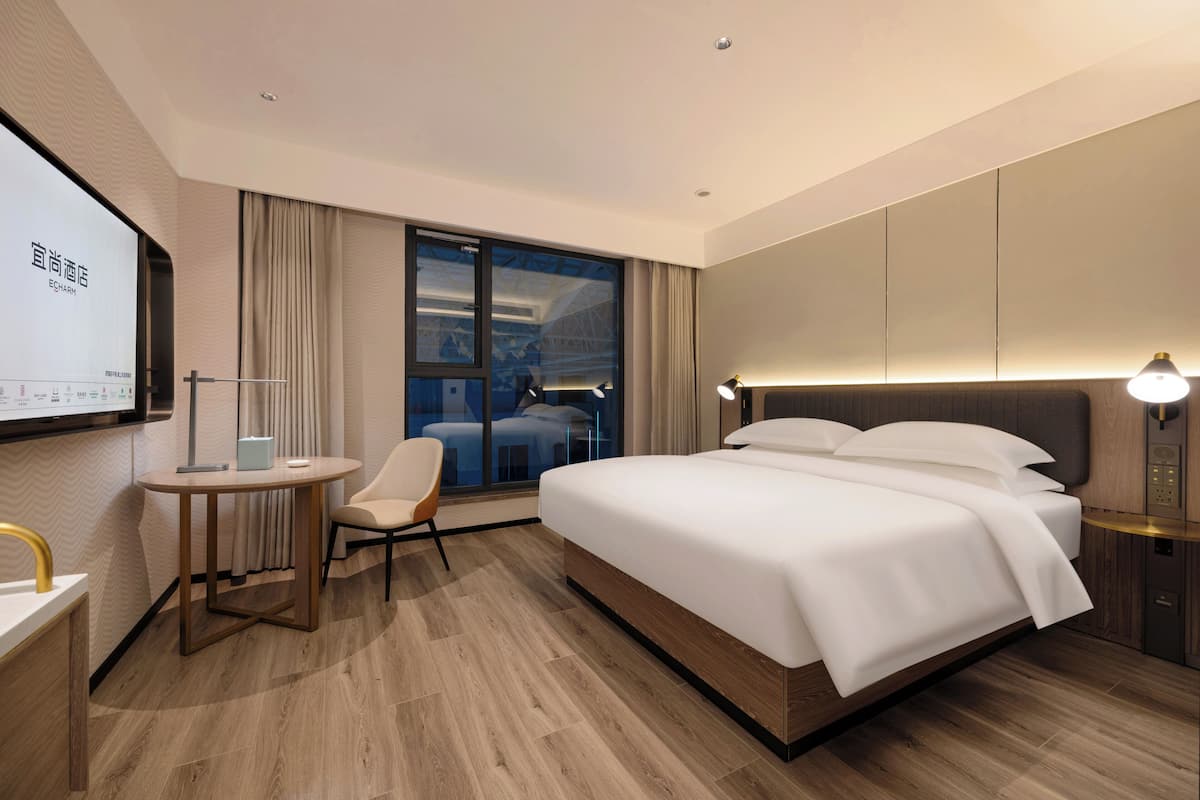 Una habitación de hotel moderna con una cama grande, un escritorio con silla, una pantalla de televisión y una ventana con cortinas.