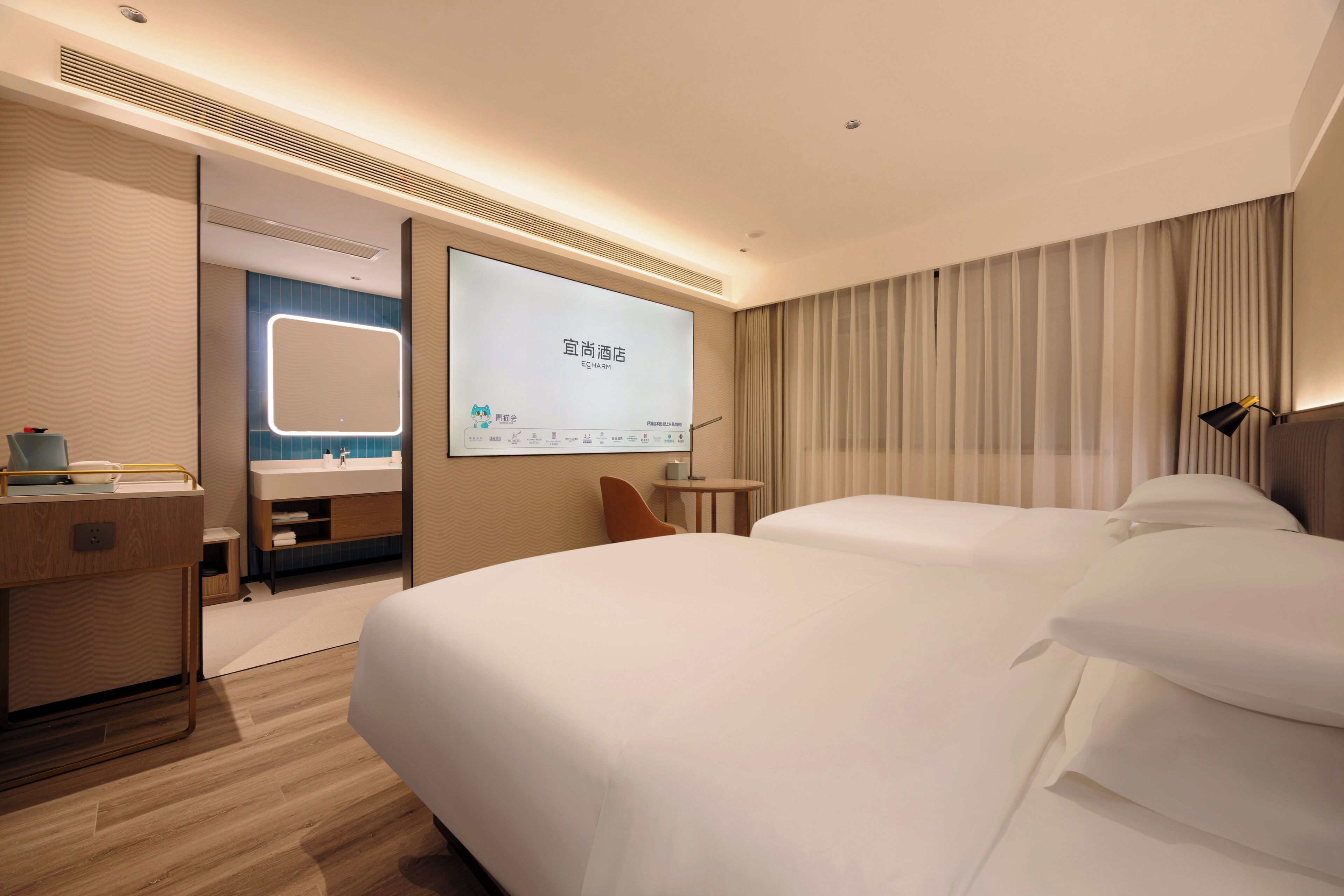 Habitación de hotel con dos camas, un espejo grande, un escritorio con una silla y una pantalla de televisión mostrando una presentación.
