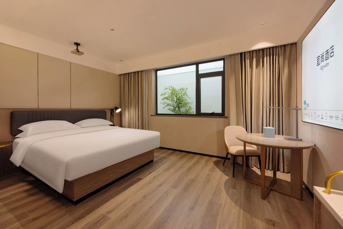 Una habitación de hotel con cama, escritorio, silla, ventana con cortinas y una pantalla montada en la pared.
