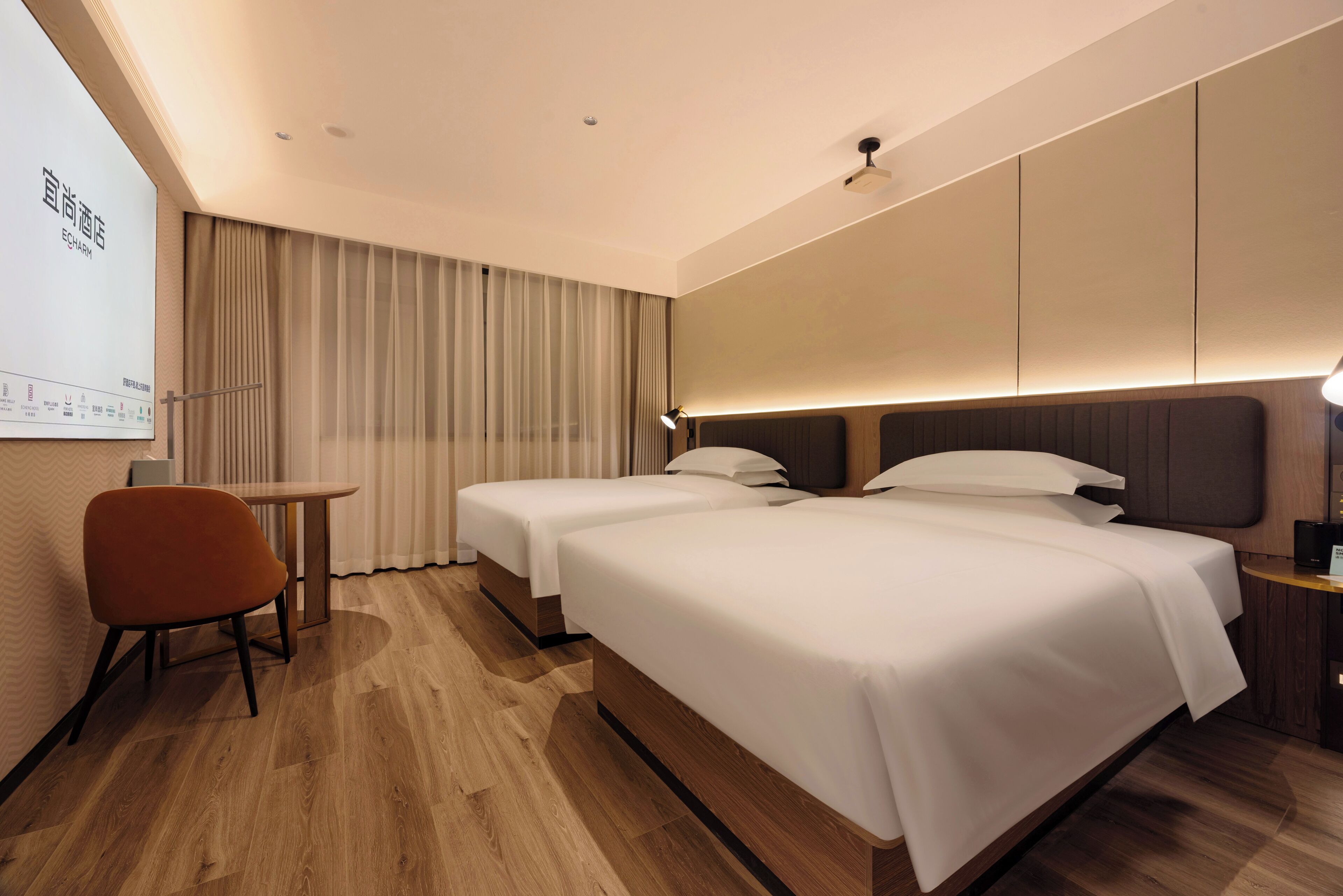 Une chambre d’hôtel avec deux lits, une petite table, une chaise et un écran de projection fixé au mur.
