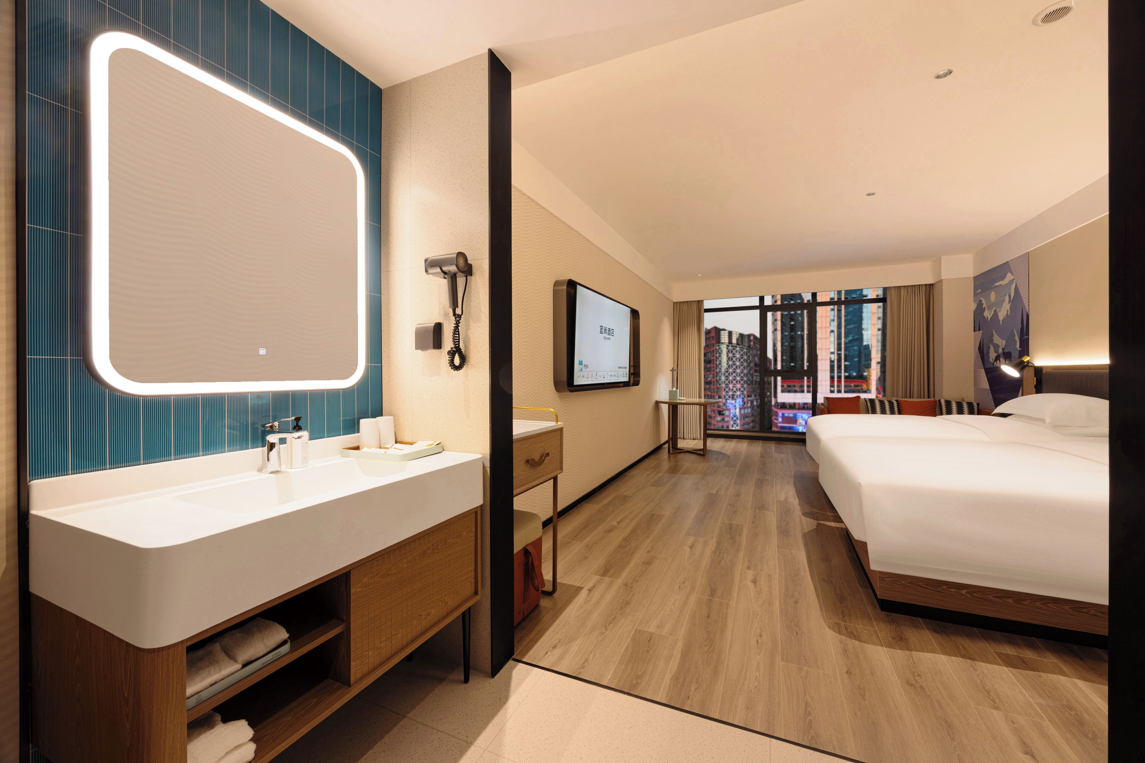 Quarto de hotel com um espelho grande, duas pias, uma cama, uma televisão e vista para edifícios pela janela.