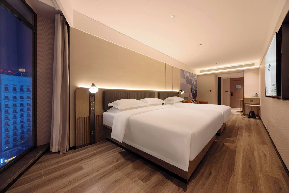 Una habitación de hotel moderna con una cama grande, una mesita de noche, un escritorio pequeño y un amplio ventanal con vistas a la ciudad.