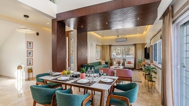 A modern dining area with a high ceiling, a central table set for four, and a cozy seating area with a sofa and chairs. - ELIVAAS Luxe Shourya Eden 3 BHK villa  (Udaipur)