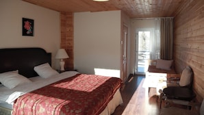 Ein gemütliches Zimmer mit Holzbalkendecke, einem Bett mit roter Bettdecke, einem Nachttisch mit Lampe, einer Holzbank und einer Tür, die nach draußen führt.