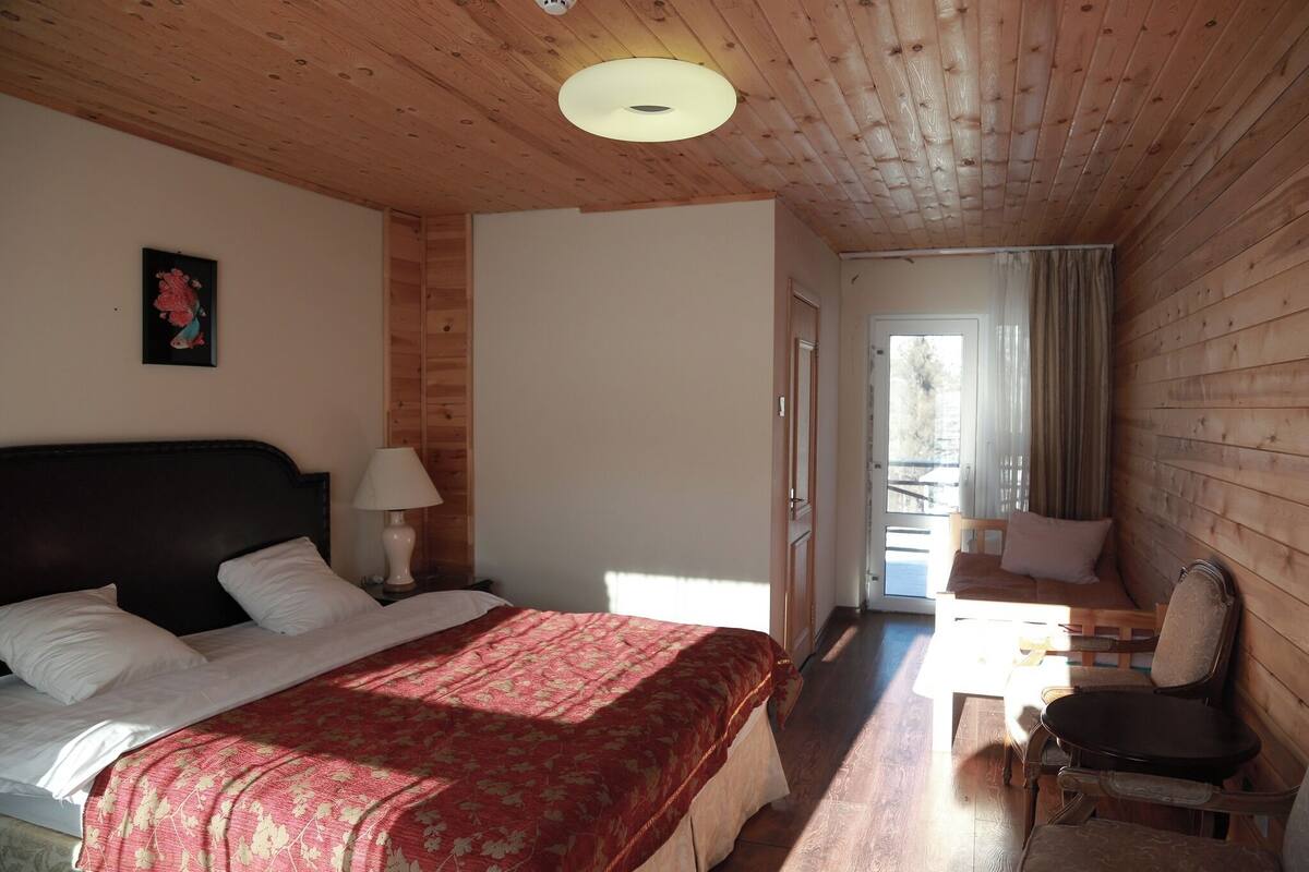 A cozy room with a wooden ceiling, a bed with a red bedspread, a nightstand with a lamp, a wooden bench, and a door leading outside.