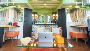 Een moderne woonkamer met een glazen salontafel, een laptop en een schaal. Er zijn twee oranje poefs, een bank met gekleurde kussens en een eettafel met stoelen.