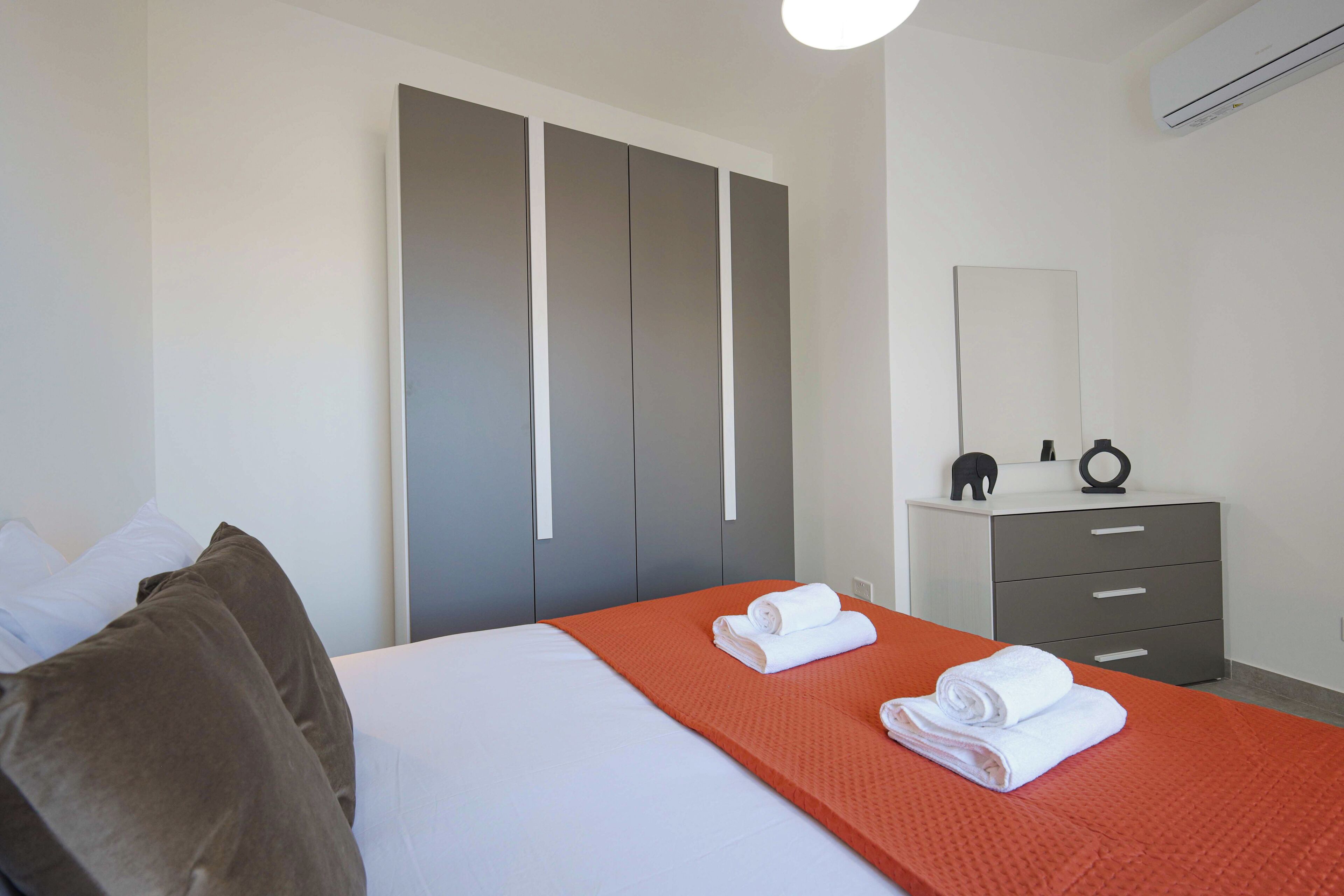 A hotel room with a bed, grey wardrobe, a small bedside table with a mirror, and a red bedspread.