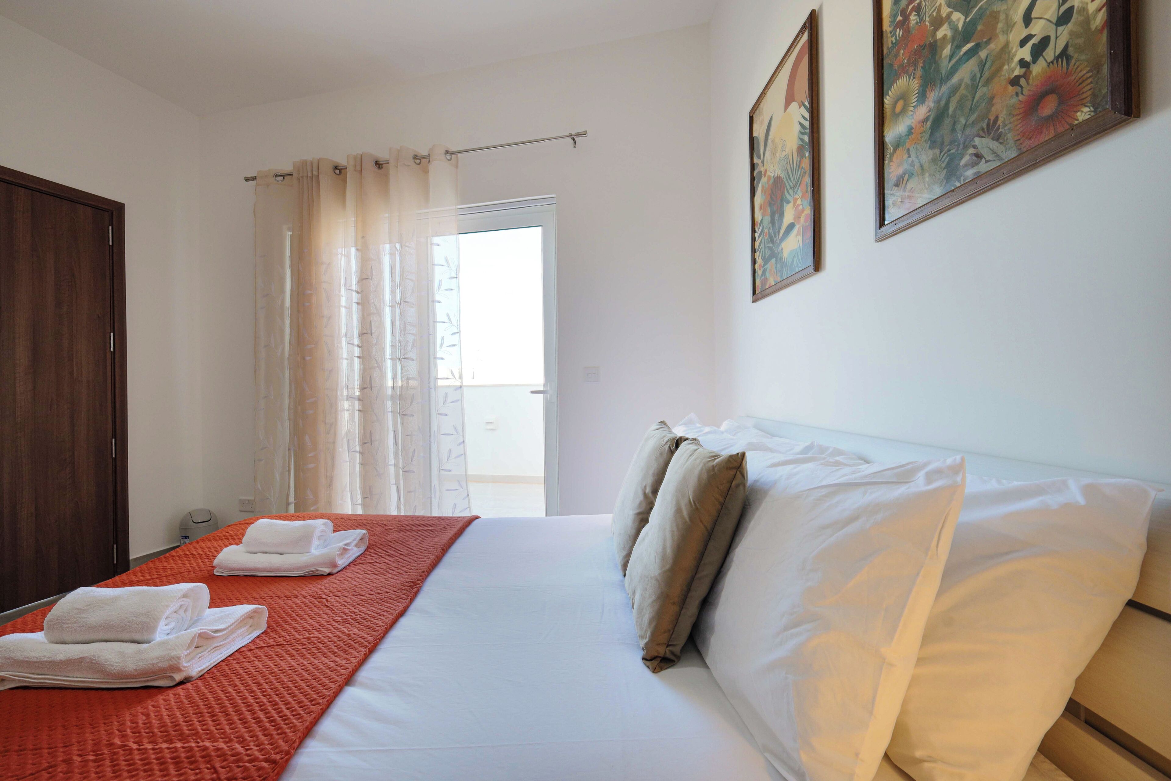 A hotel room with a bed, a window with curtains, a door, and two paintings on the wall.
