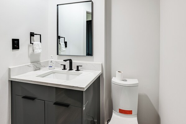 Een moderne badkamer met een witte wastafel, een zwarte kraan en een grote spiegel. Op het aanrecht staan een toiletrolhouder en een zeepdispenser.