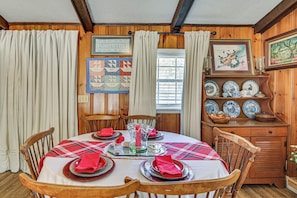 Un comedor con una mesa redonda preparada para cuatro personas, sillas de madera, mantel a cuadros y un aparador con platos decorativos.