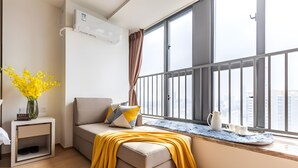 A modern living room with a sofa, a small side table with a vase, a window with a view of buildings, and a balcony railing. - Yazhi Apartment (Guangzhou)