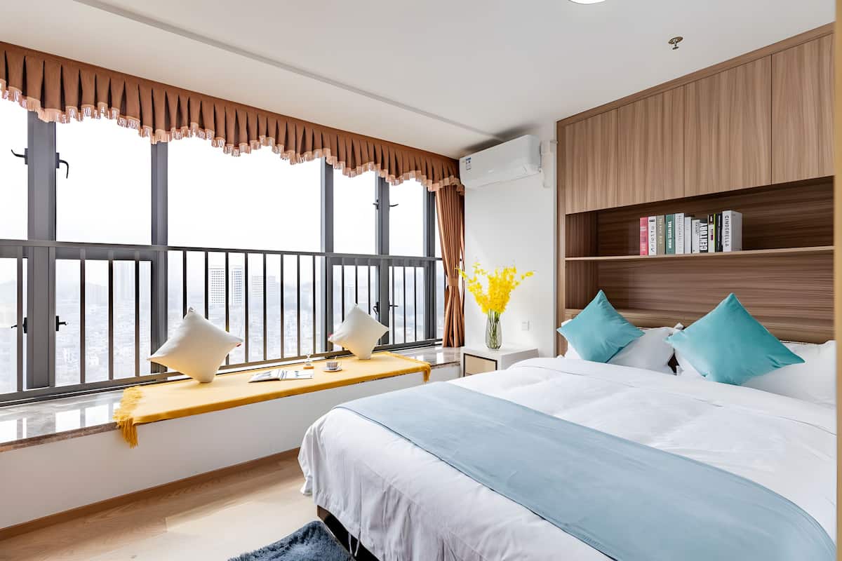 Una habitación de hotel moderna con una cama grande, un asiento junto a la ventana, una mesita de noche con lámpara, una estantería y vistas a la ciudad.