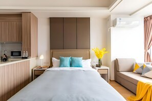 A modern hotel room with a large bed, a bedside table with a lamp, a small desk, a sofa, and a kitchen area with a microwave. - Yazhi Apartment (Guangzhou)