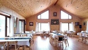 Une salle à manger spacieuse avec de hauts plafonds en bois et de grandes fenêtres. Les tables sont dressées avec des nappes blanches et des chaises en bois.