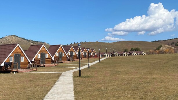 Une rangée de cabanes en bois, chacune avec une porte numérotée, un ciel dégagé et un champ verdoyant avec des collines en arrière-plan.