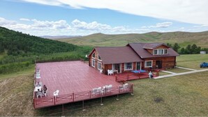 Une cabane en bois avec une terrasse rouge et un porche, entourée d’un paysage verdoyant et de collines.