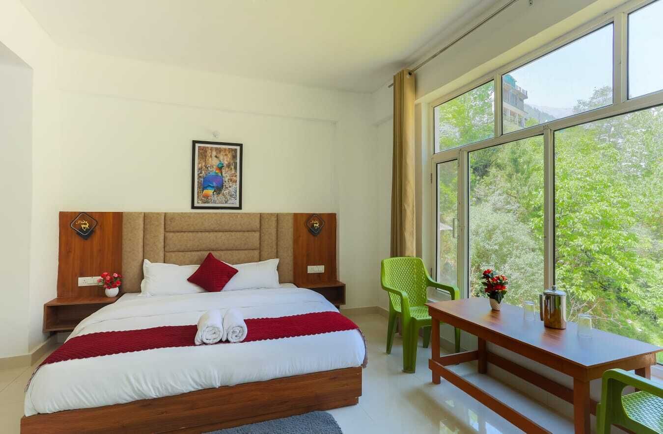 A bedroom with a large bed, a wooden headboard, a nightstand, a green chair, a wooden table, and a window with a view of greenery.