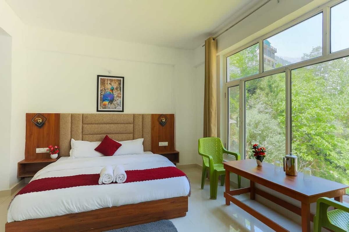 A bedroom with a large bed, a wooden headboard, a nightstand, a green chair, a wooden table, and a window with a view of greenery.