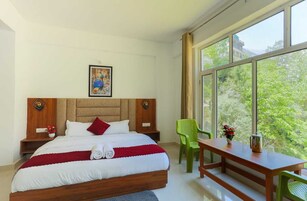 A bedroom with a large bed, a wooden headboard, a nightstand, a green chair, a wooden table, and a window with a view of greenery.