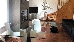 A modern living room with a glass dining table, white chairs, a black sofa, a TV, and a wooden staircase. - Gîte les Coquelicots facing the river (Vignonet)