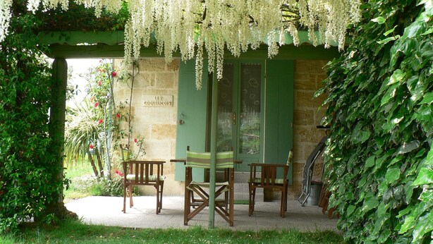 A covered outdoor seating area with wooden chairs and a table, surrounded by greenery and a stone wall. - Gîte les Coquelicots facing the river (Vignonet)
