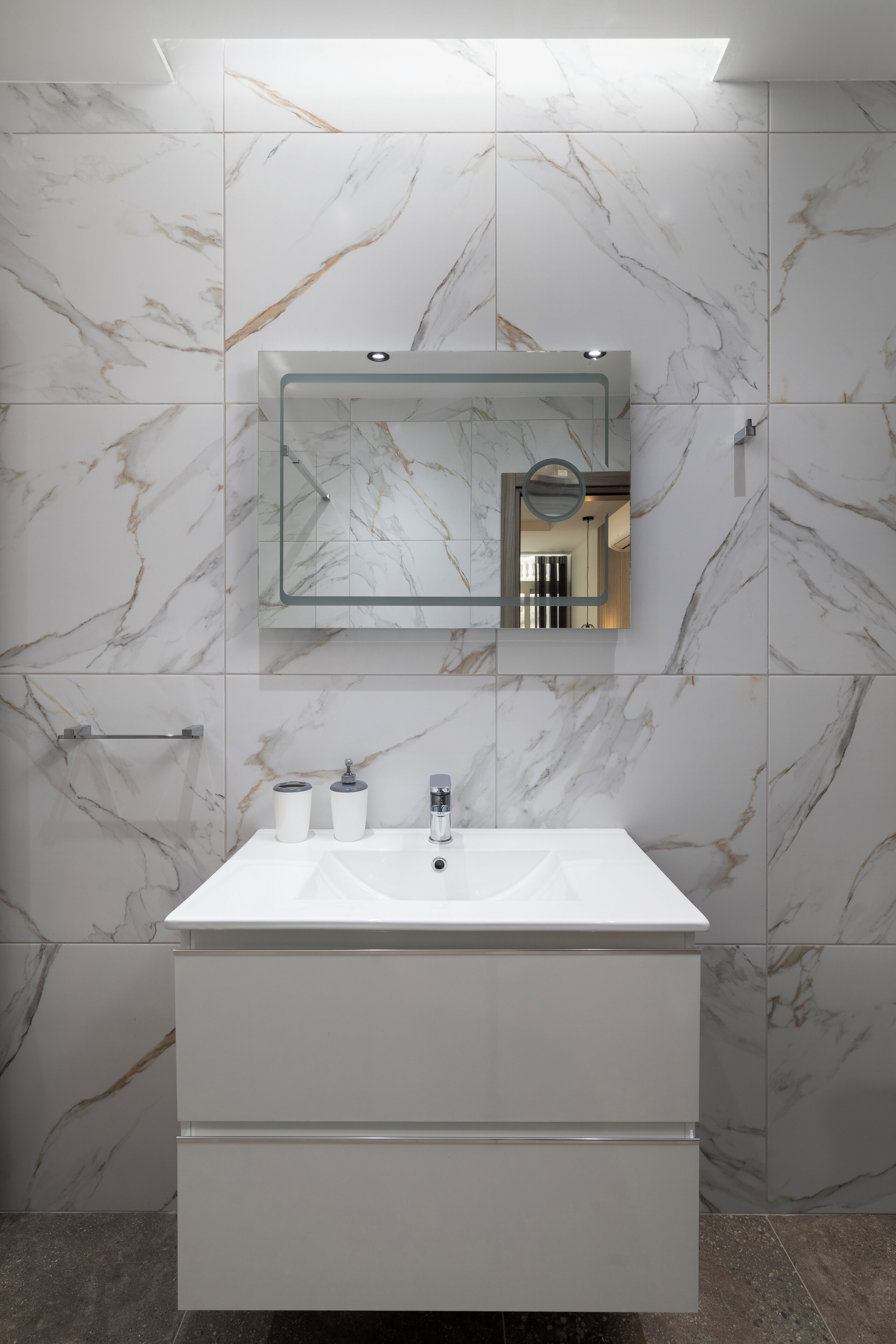 A bathroom with a marble wall, a white sink, a mirror, and a towel rack.