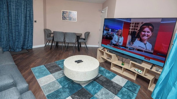 Een woonkamer met een televisie, een rond poefje, een eettafel met stoelen en een blauw gordijn.
