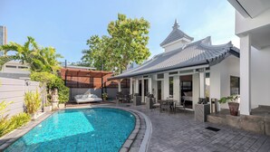 Een zwembad met betegeld zonnedek, een overdekt terras met tuinmeubilair en een gebouw in traditionele stijl met een betegeld dak.