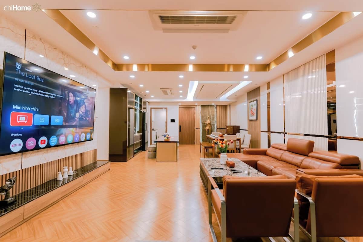 A modern living room with a flat-screen TV, wooden flooring, a brown leather sofa, and a dining area with a marble table.