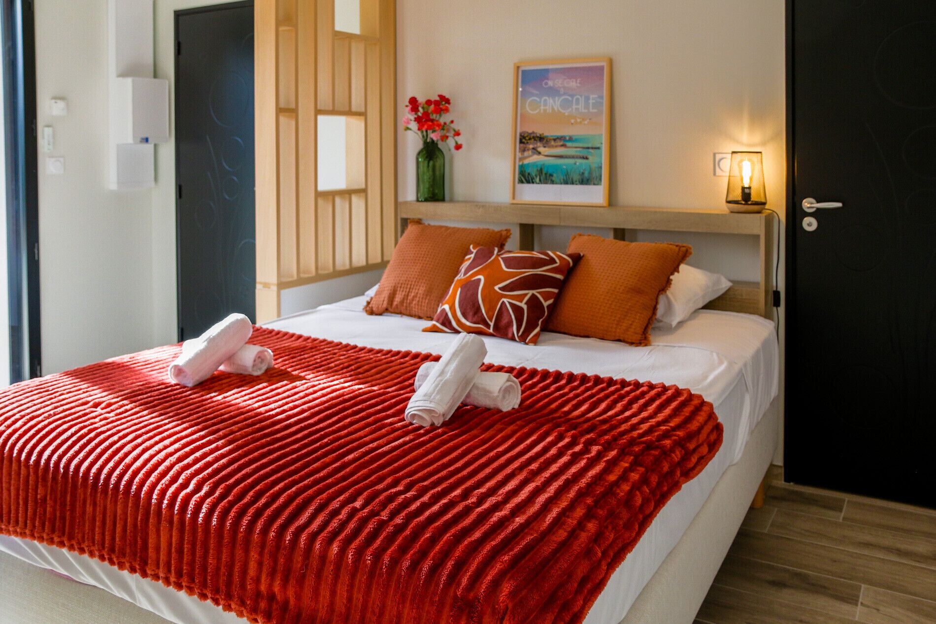 A hotel room with a bed, a red textured blanket, two rolled towels, a wooden headboard, a framed picture, and a bedside lamp.
