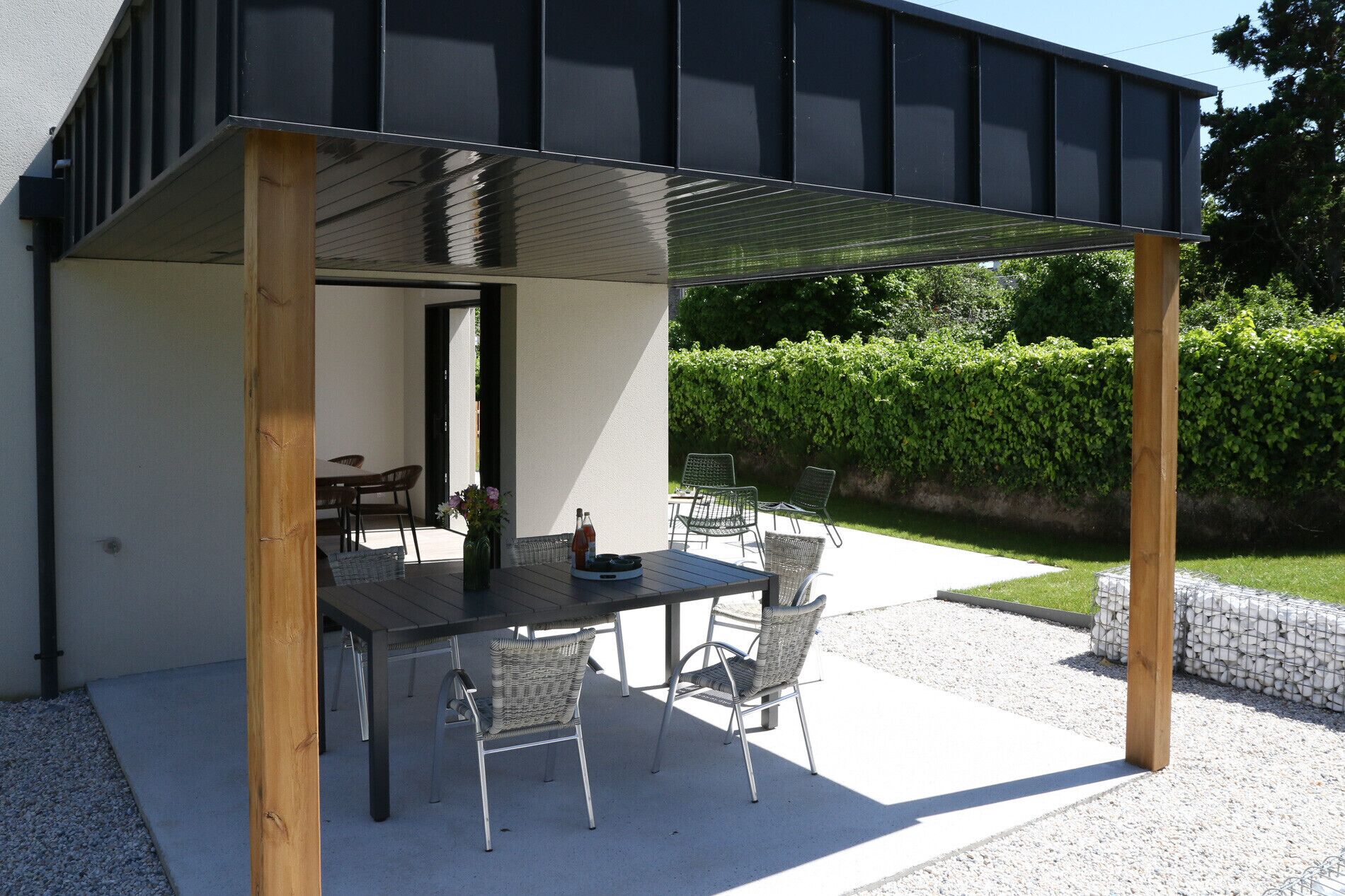 Eine Terrasse im Freien mit Tisch und Stühlen, einer Holz-Pergola und einer Hecke im Hintergrund.