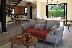 Ein modernes Wohnzimmer mit einer Couch, einem Couchtisch mit Flasche und Gläsern, einem Essbereich mit Tisch für Zwei und einer Küche im Hintergrund.