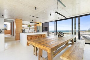 A modern kitchen with a central island, wooden dining table, and large windows offering a view of the ocean. - Nowhere Els (Rooiels)