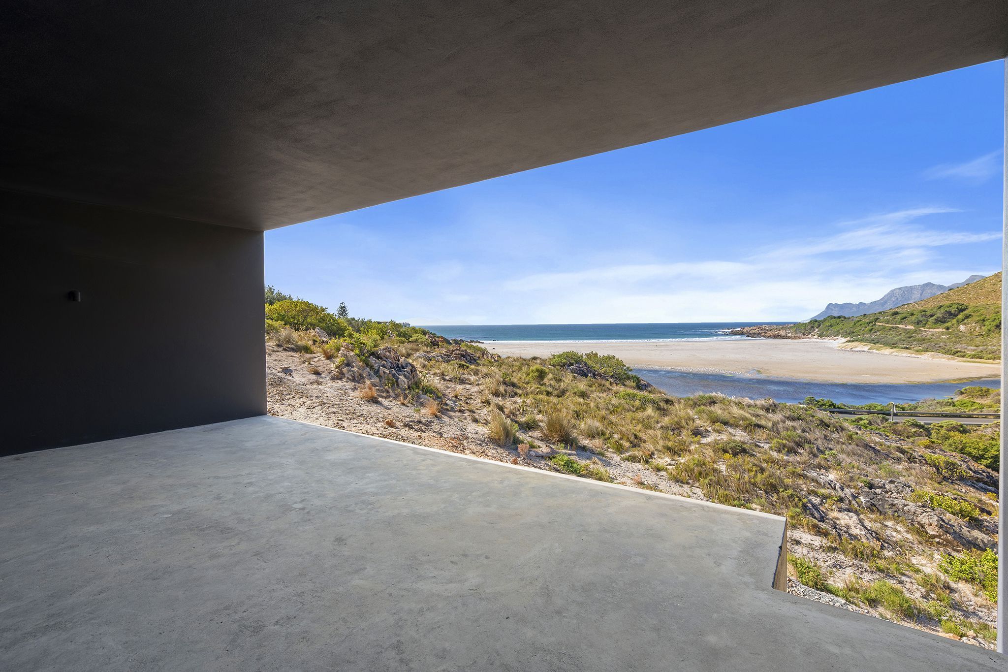 Un espace intérieur en béton donnant sur une plage et des montagnes.