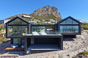 Una casa moderna de dos pisos amb grans finestres de vidre, un balcó i vista a una muntanya.